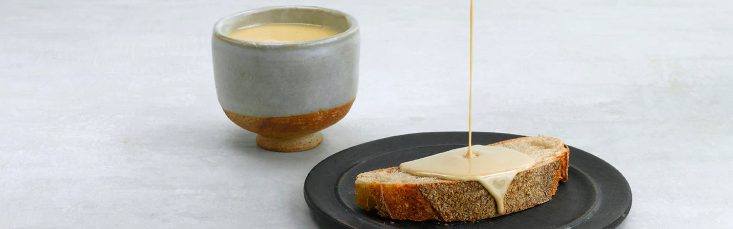 A piece of toast with tahini being drizzled onto it and a cup of tahini on the side.