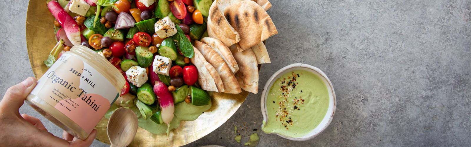 Salad bowl with Tahini Sauce and a hand pouring a jar of tahini over top.