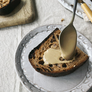 A slice of toast on a small plate with a big spoon full of tahini being spread onto it.