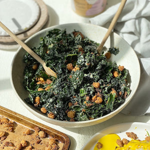 A kale caesar salad in a bowl, sprinkled with chickpeas. There is a pan of chickpeas on the side, a plate of cauliflower and assorted linens and plates.