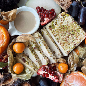 A sliced piece of pistachio sits on a cutting board surrounded by jewel-toned fruits and bread, and a small cup of tahini.