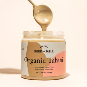 A jar of tahini with a spoon dripping tahini into it from above.