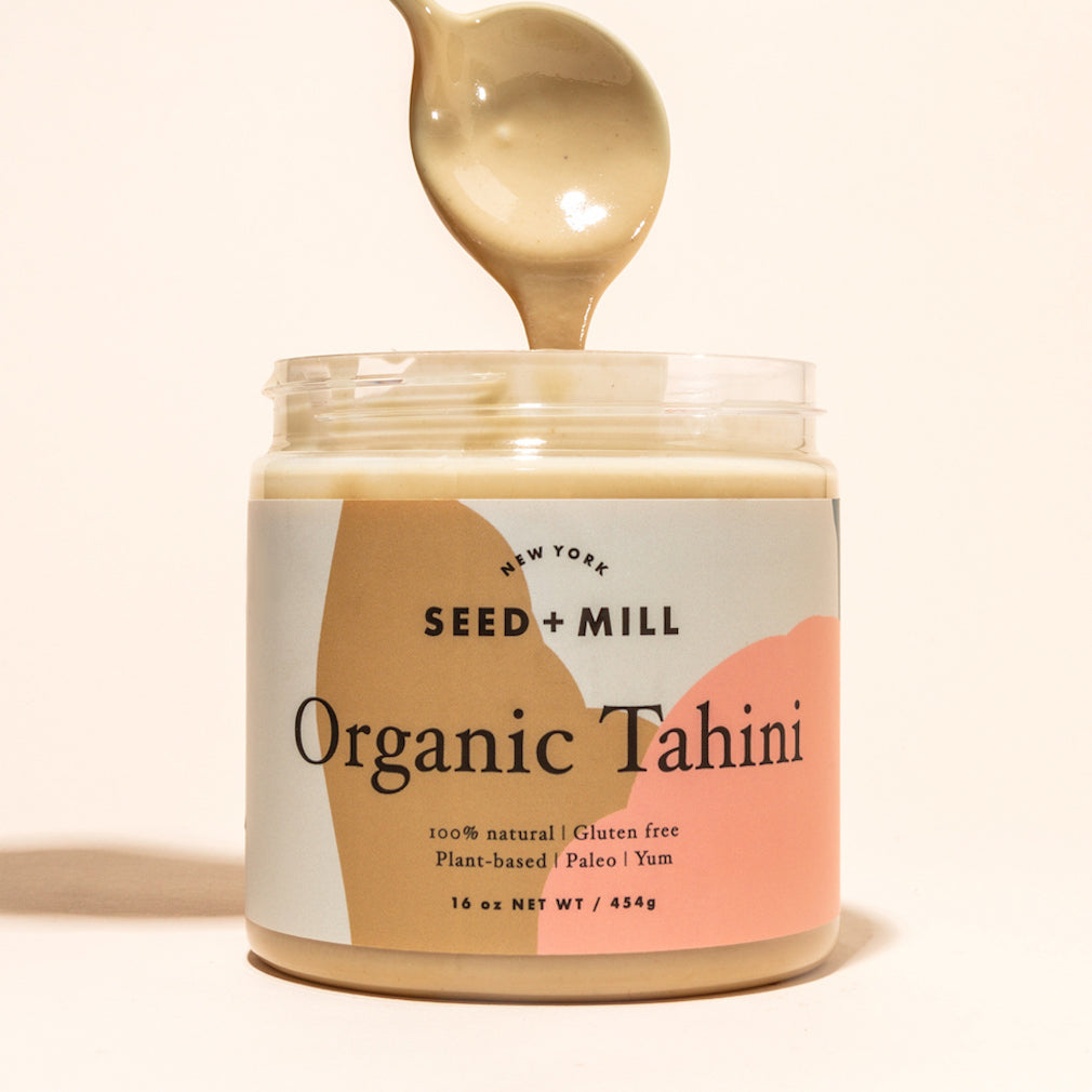 A jar of organic tahini with a spoon full of tahini dripping into it.