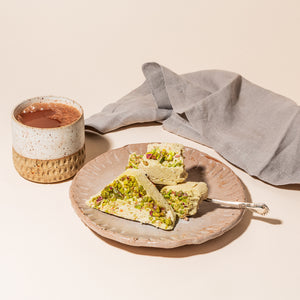 A plate with wedges of pistachio halva and a fork on top. There is a napkin on one side of the plate and a mug of cocoa on the other.