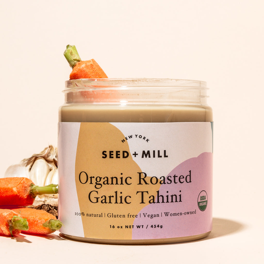 A jar of roasted garlic tahini with carrots and garlic on the side, and a carrot dipped into the jar.