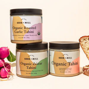 An image of three tahini jars, two are stacked on top of one another. On one side is a bunch of radishes and on the other, slices of bread.