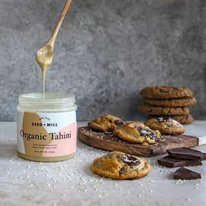 A cutting board covered in tahini chocolate chip cookies, sprinkled with sesame seeds, next to a small pile of dark chocolate and a jar of organic tahini with a spoon full of it dripping above.