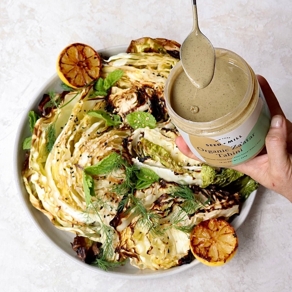 A platter of charred cabbage topped with herbs and lemon wedges. A hand holds a jar of the za'atar tahini over the platter and is spooning it onto the cabbage.