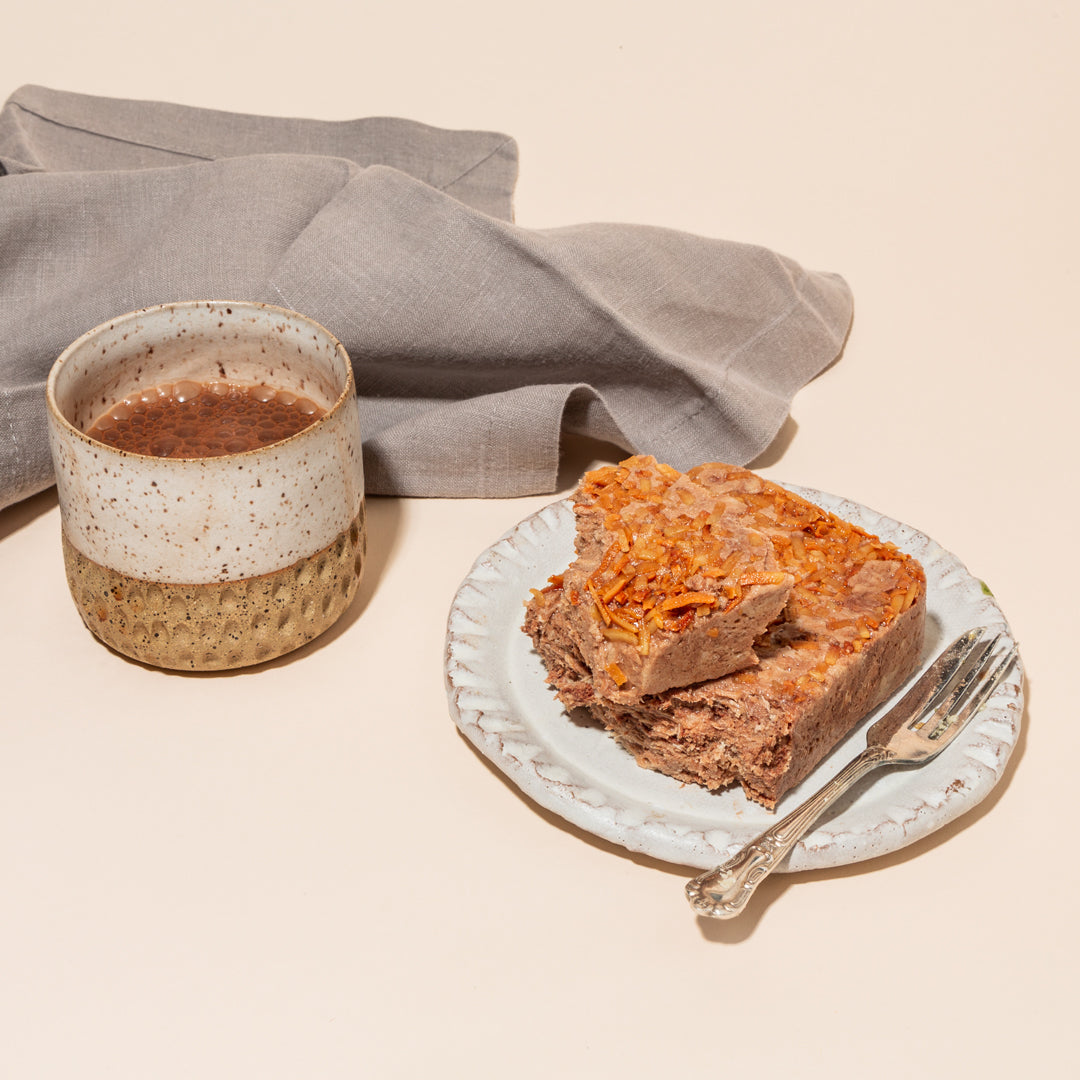 A plate with wedges of dark chocolate and toasted coconut halva and a fork on top. There is a napkin on one side of the plate and a mug of cocoa on the other.