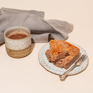 A plate with wedges of dark chocolate and toasted coconut halva and a fork on top. There is a napkin on one side of the plate and a mug of cocoa on the other.