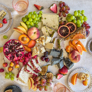 A snack board covered in bright fruits, nuts, slices of halva with an assortment of plates and glassware surrounding it. 