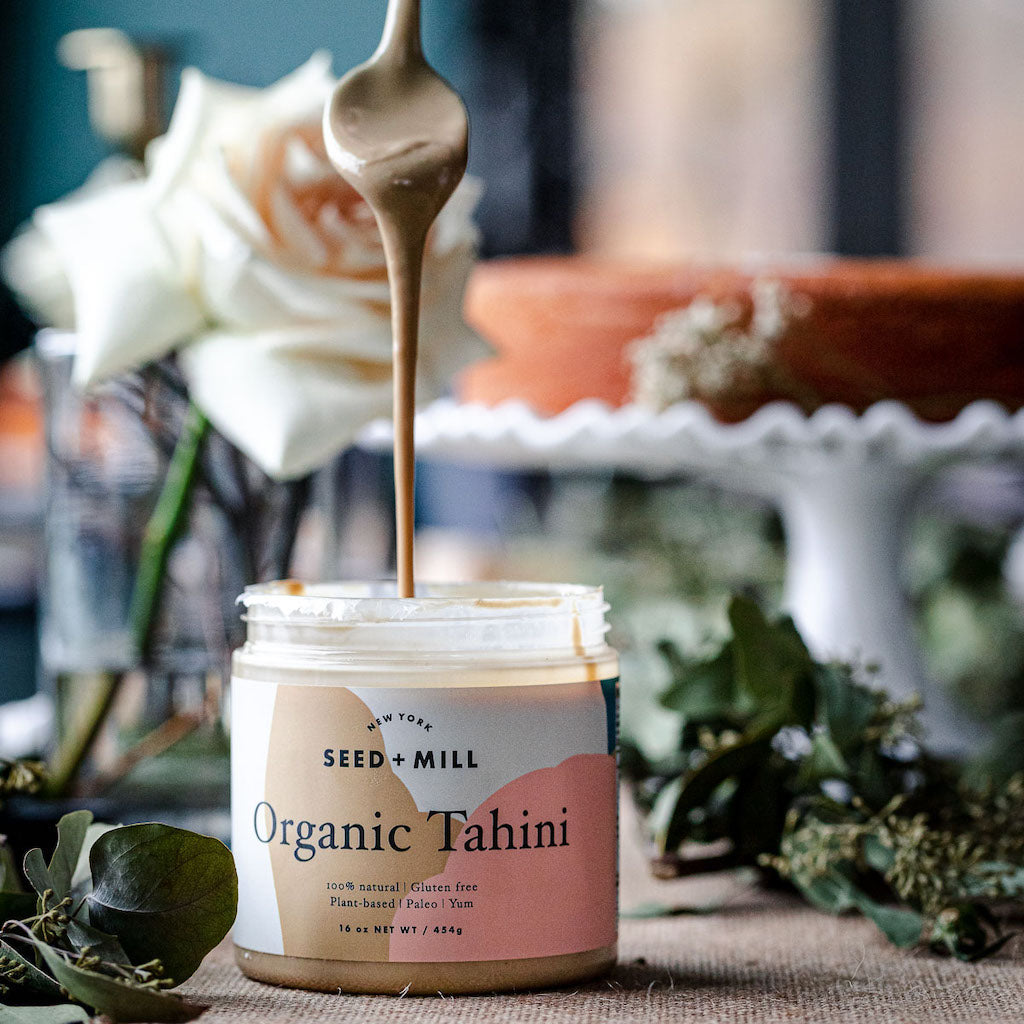 A jar of tahini surrounded by abstracted florals, with a spoon drizzling tahini into the jar.