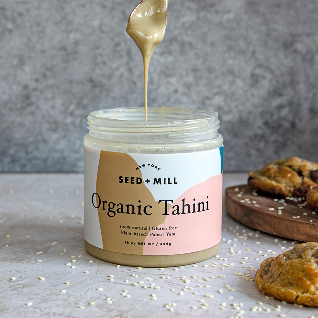 A jar of tahini next to a pile of cookies with a spoon over top, dripping tahini from above. 